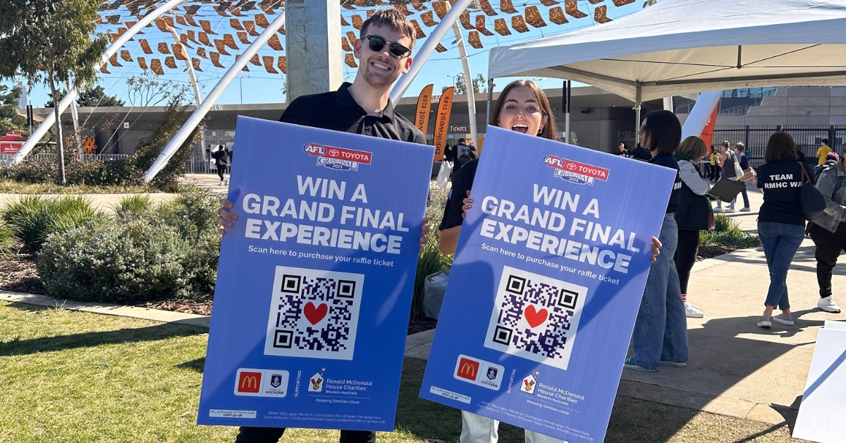 2 people standing in front of Optus Stadium holding large signs that say 'WIN A GRAND FINAL EXPERIENCE'